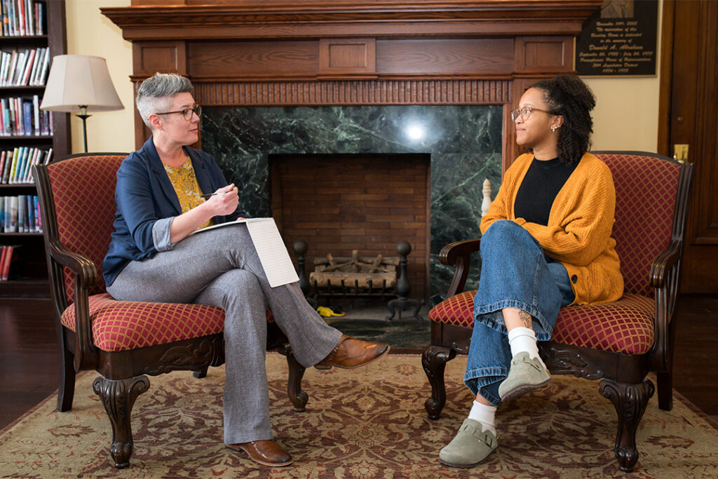 Two professionals sit across from one another in front of a fireplace, one with notepad and pen in hand.
