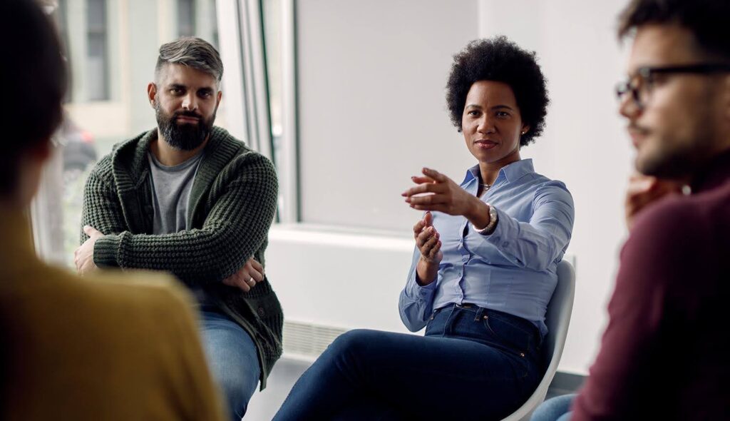 A group is seated in chairs arranged in a circle; a woman gestures while she speaks to the group, who listen attentively.