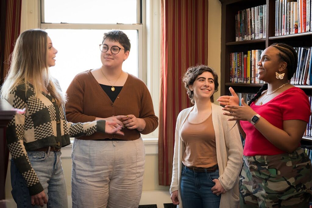 Four students stand and chat in a cozy room with bookshelves and a window, smiling and engaged in friendly conversation.