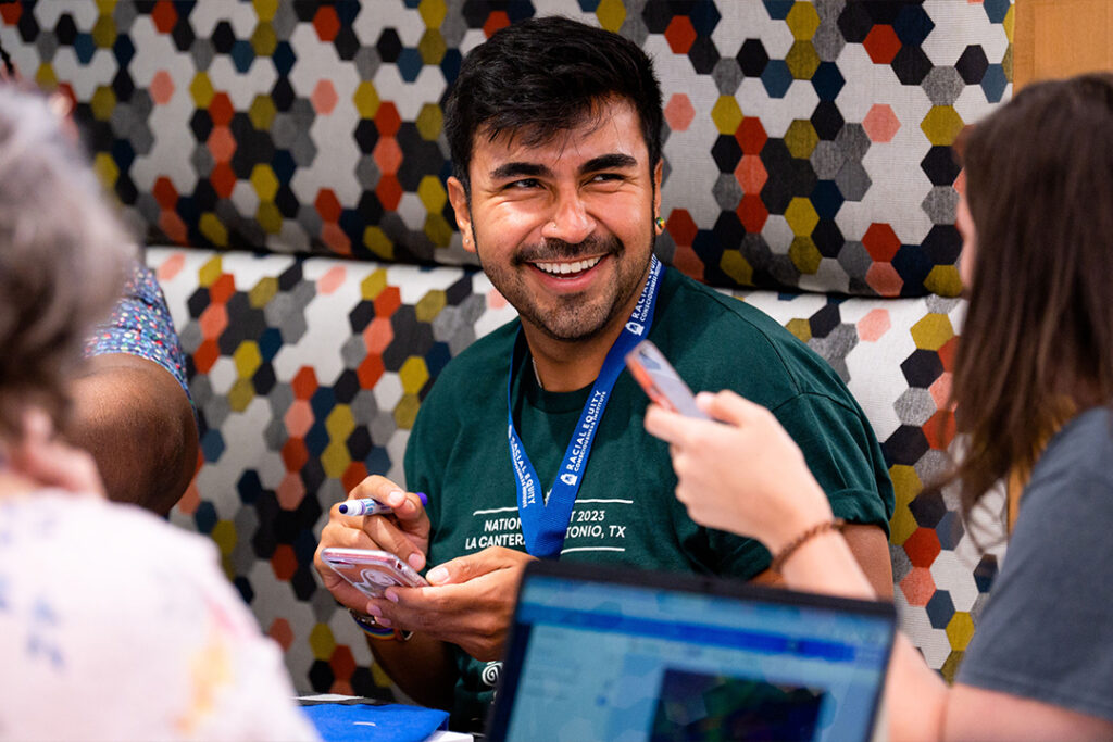 A graduate student wearing a lanyard holds his phone and a marker while smiling in a group of other adults.