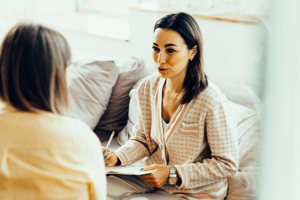 A woman holding a pen and notebook speaks with another woman during a seated, informal conversation in a cozy indoor setting.